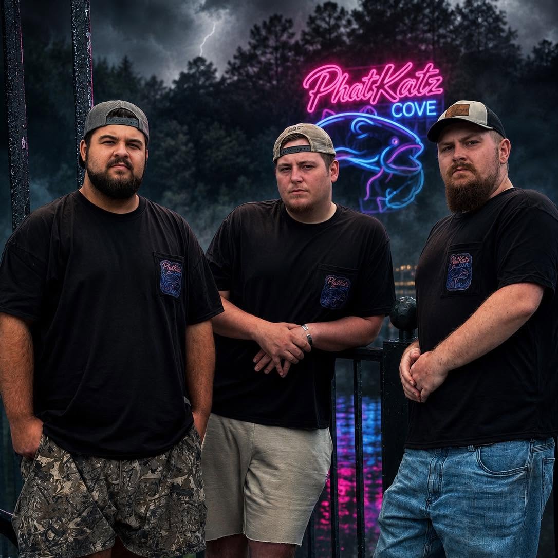 Three PhatKatz Cove crew members wearing black logo shirts beside the water, with the blue and pink neon Cove sign behind them